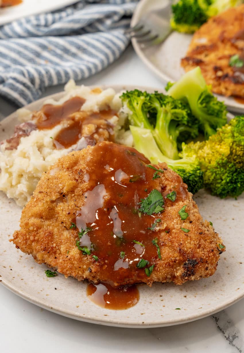 pork schnitzel topped with brown gravy on a white plate with steamed broccoli and mashed potatoes on the side