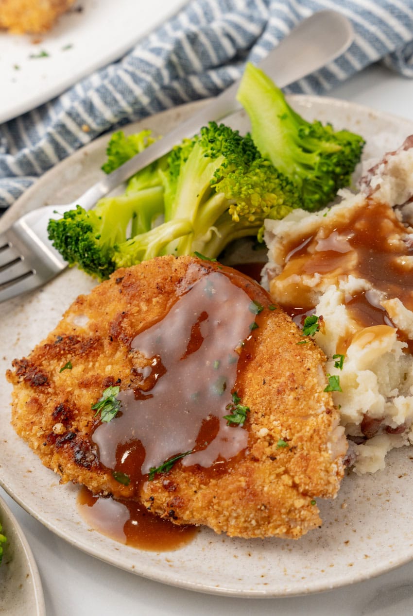 pork schnitzel topped with brown gravy on a white plate with steamed broccoli and mashed potatoes on the side