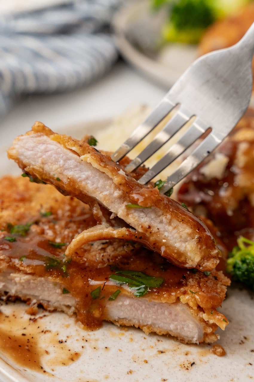 a silver fork lifting a piece of pork schnitzel covered in brown gravy off a white dinner plate