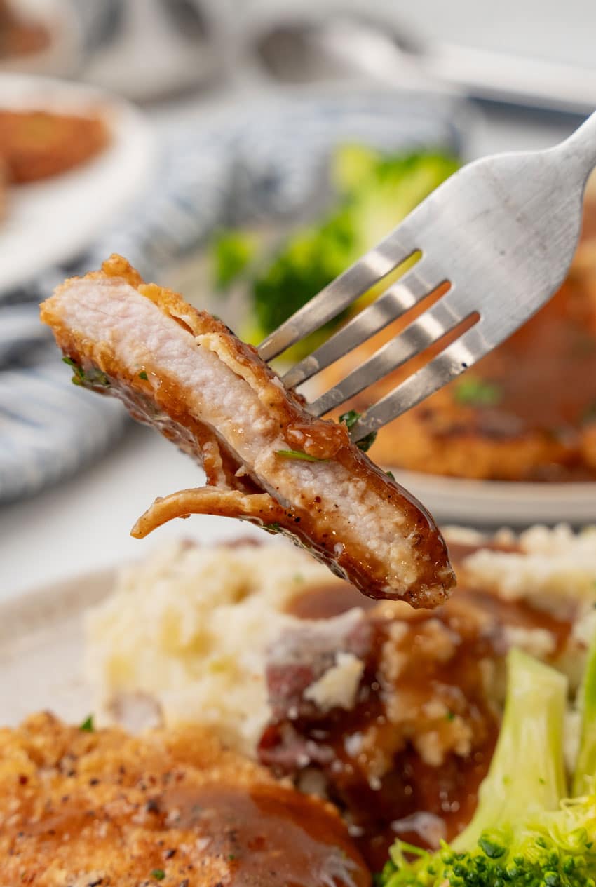 a silver fork lifting a piece of pork schnitzel covered in brown gravy off a white dinner plate