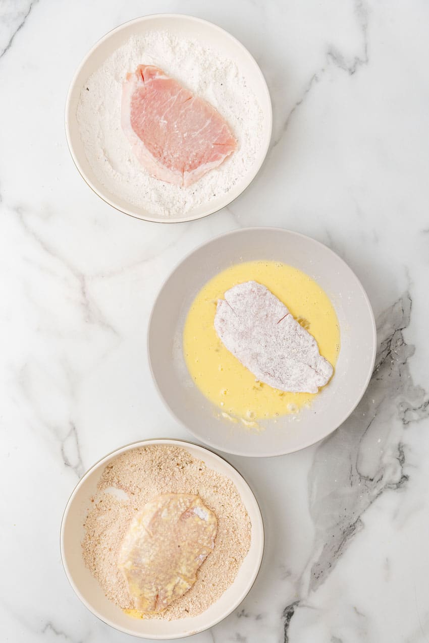 pork chops being dredged in three bowls filled with flour, beaten egg, and bread crumbs