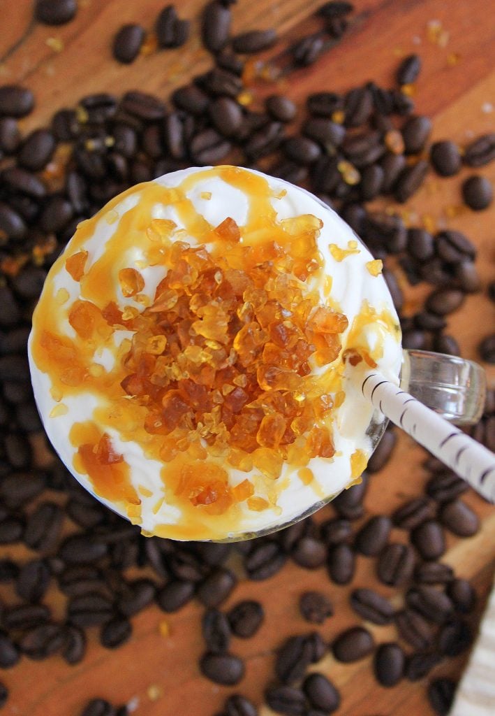 Seen from overhead Cold Creme Brulee Coffee with Caramel in a glass mug is surrounded by coffee beans on a brown cutting board. The creamy coffee's topped with whipped cream with crunchy caramel bits and a drizzle of caramel sauce. A straw sticks out from the mug