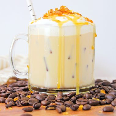 Cold Creme Brulee Coffee with Caramel in a glass mug surrounded by coffee beans on a brown cutting board. The creamy coffee's topped with whipped cream with crunchy caramel bits and a drizzle of caramel sauce. A straw sticks out from the mug