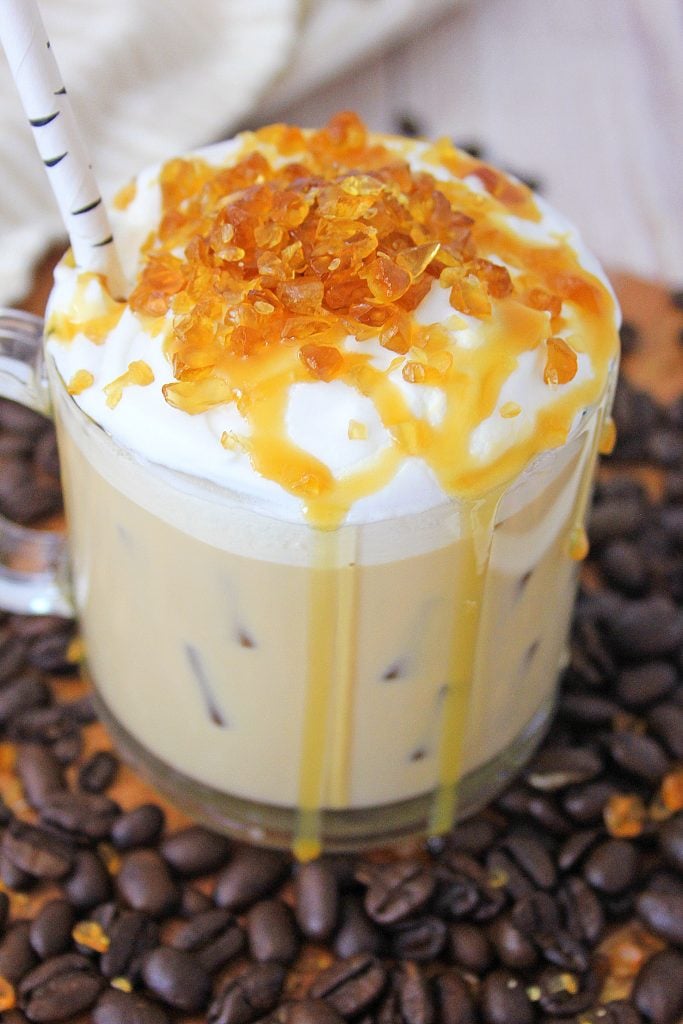 Cold Creme Brulee Coffee with Caramel in a glass mug surrounded by coffee beans on a brown cutting board. The creamy coffee's topped with whipped cream with crunchy caramel bits and a drizzle of caramel sauce. A straw sticks out from the mug