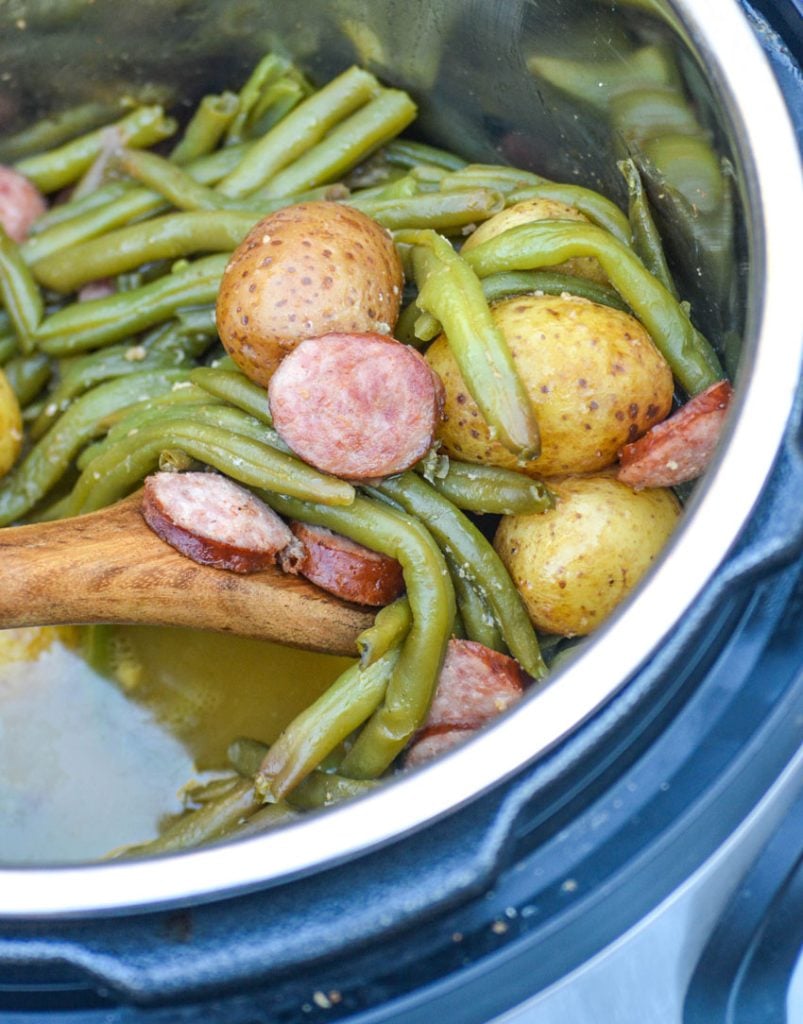 green beans, whole little potatoes, and thinly sliced smoked sausage in a pressure cooker after cooking being scooped up by a wooden spoon