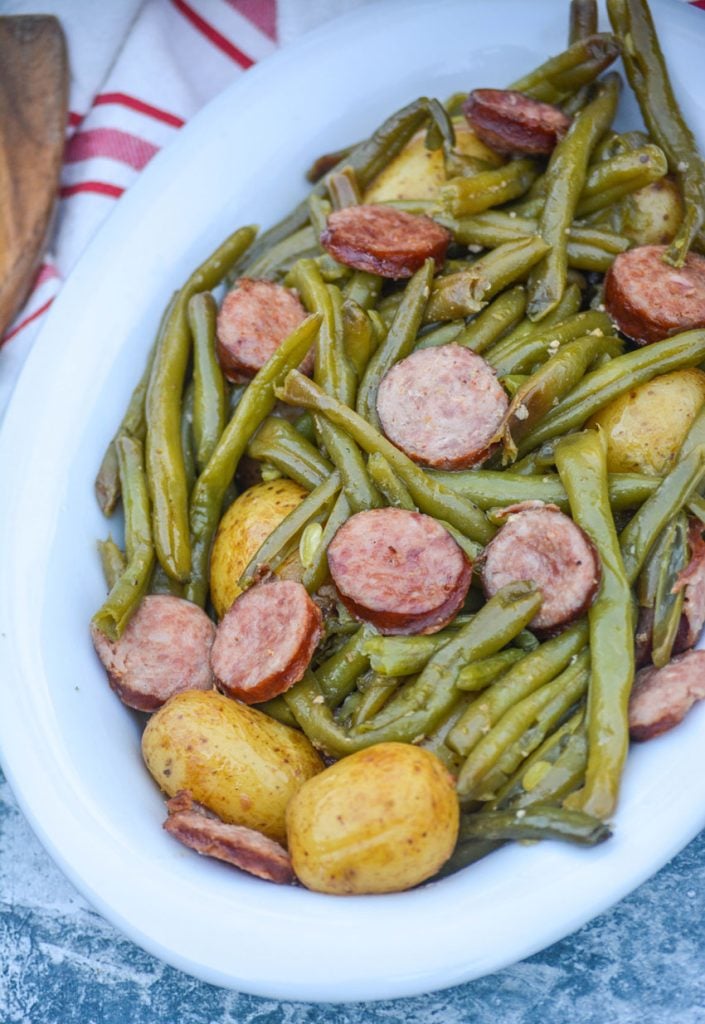 green beans, whole little potatoes, and thinly sliced smoked sausage sitting in an oval shaped white serving bowl with a red and white stripped dish towel in the background