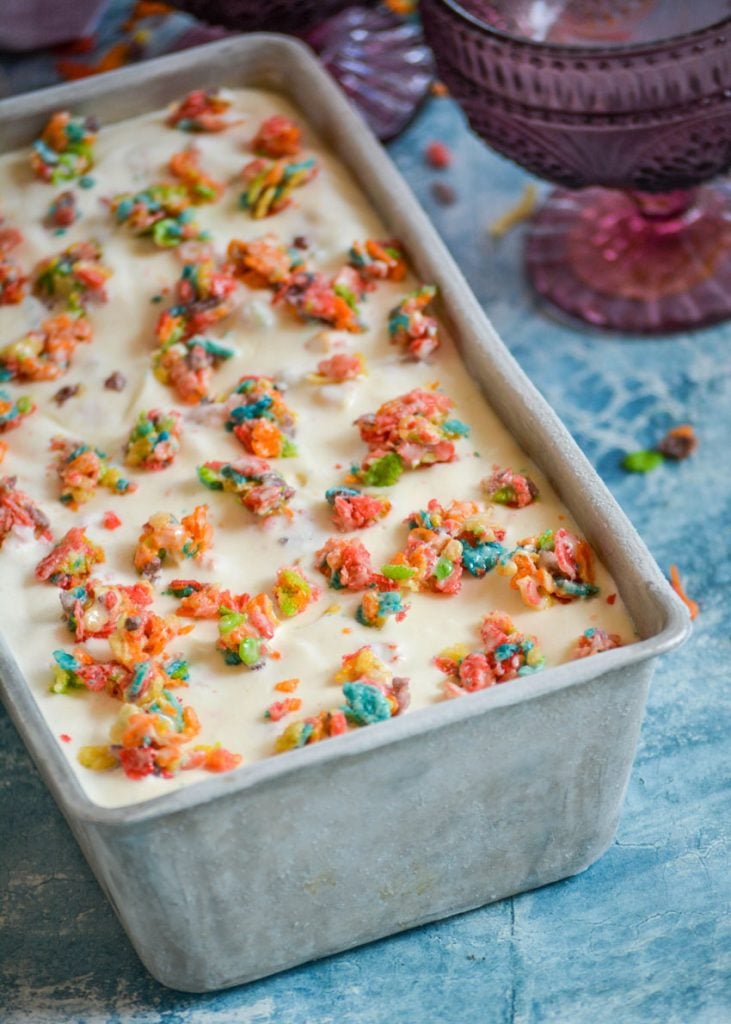 no churn fruity pebble ice cream topped with cereal clusters in a metal loaf pan on a blue background