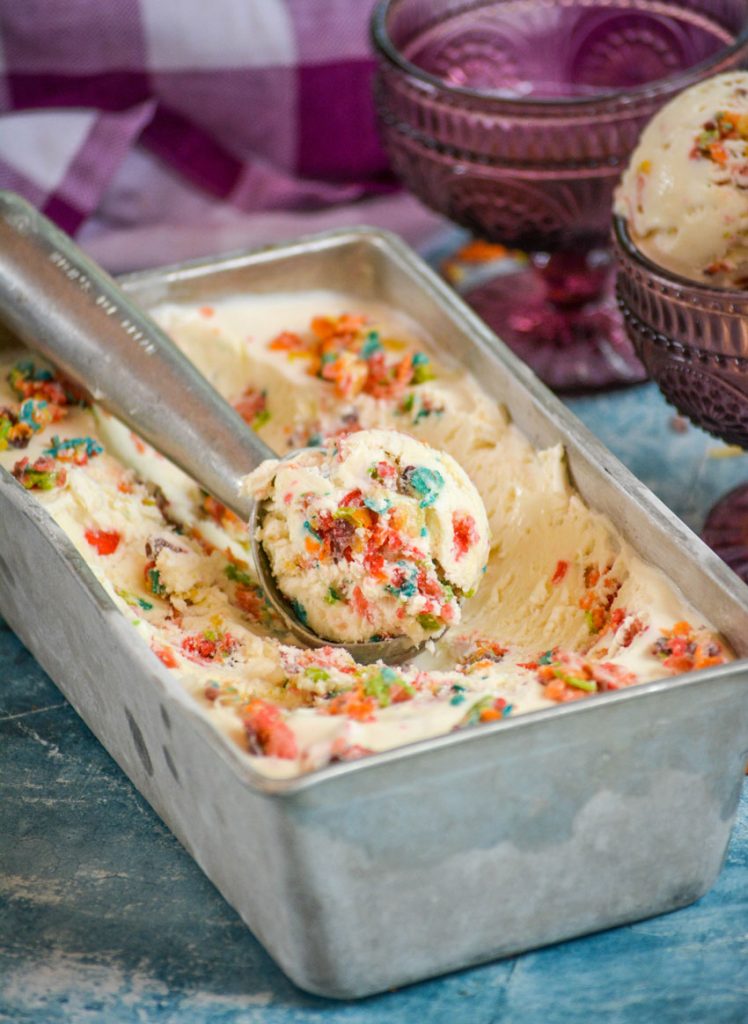 a metal ice cream scooper shown with a scoop of fruity pebble ice cream in a metal loaf pan