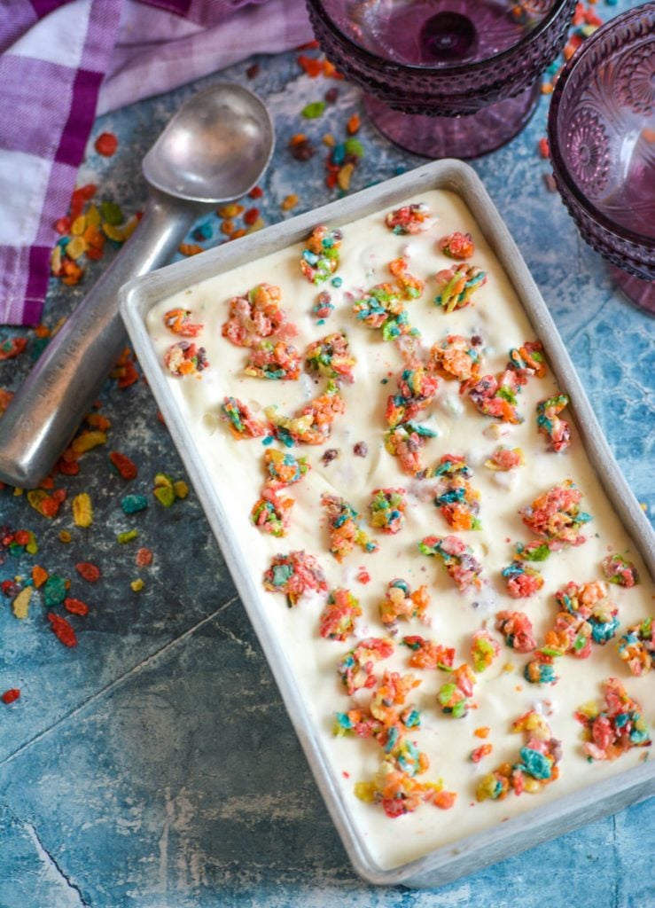 fruity pebble ice cream topped with marshmallow cereal clusters in a metal loaf pan on a blue background