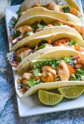 bang bang shrimp tacos lined up on a rectangular white serving platter with fresh lime wedges