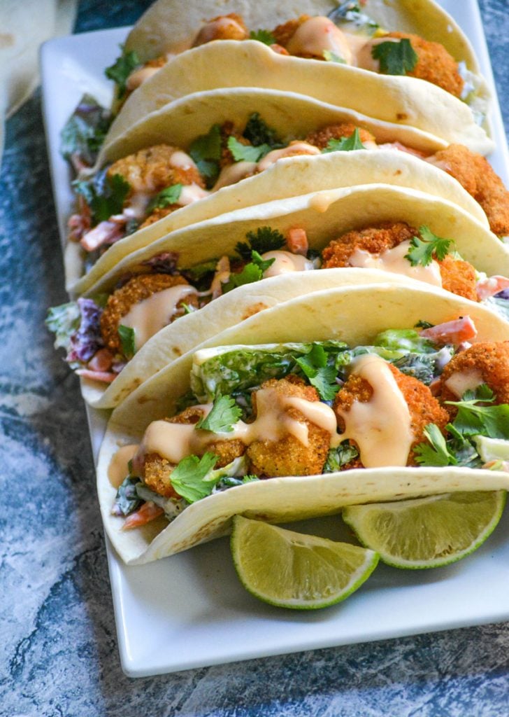 bang bang shrimp tacos lined up on a rectangular white serving platter with fresh lime wedges