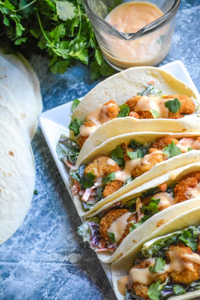 bang bang shrimp tacos lined up on a rectangular white serving platter on a blue background with extra sauce, a bunch of fresh cilantro, and flour tortillas in the background