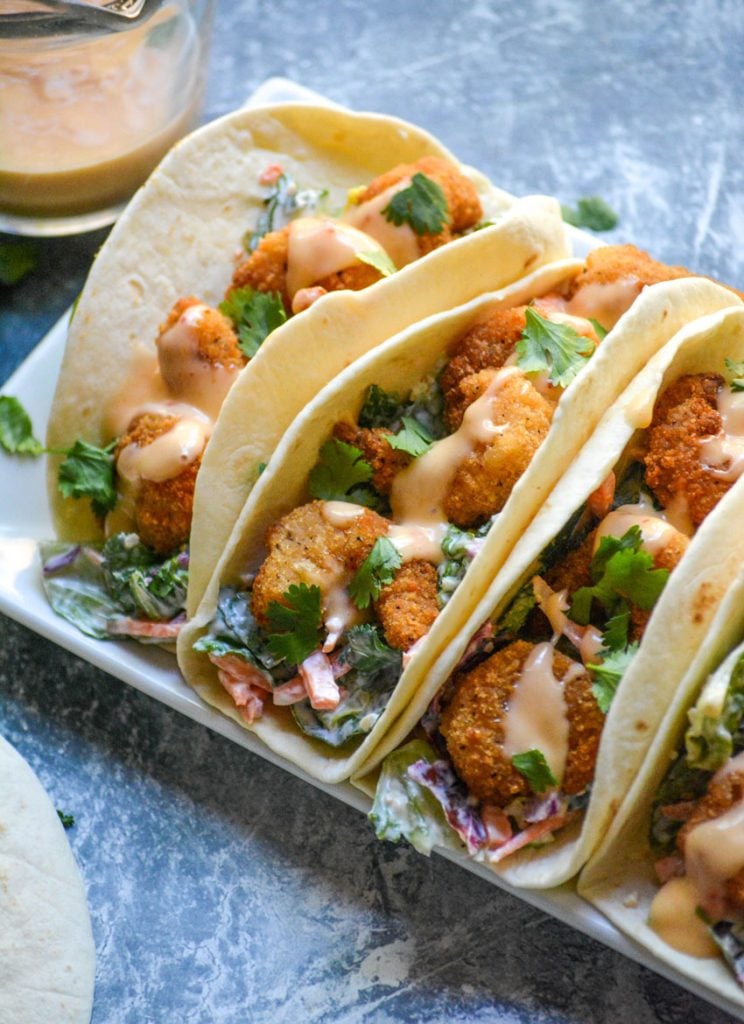 bang bang shrimp tacos lined up on a rectangular white serving platter on a blue background with extra sauce, a bunch of fresh cilantro, and flour tortillas in the background