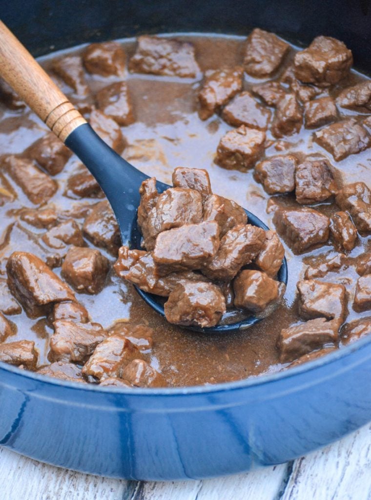 beef stew meat in brown gravy shown on a wooden spoon held above a blue pot