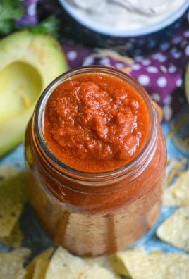 homemade taco sauce in a glass jar surrounded by tortillas chips with half a ripe avocado and a bowl of sour cream in the background