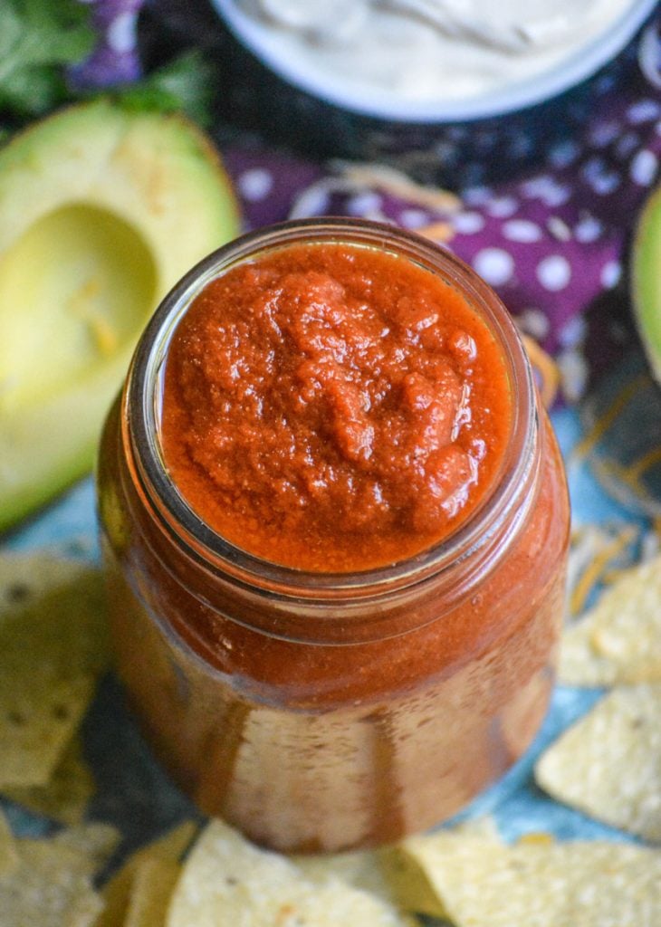 homemade taco sauce in a glass jar surrounded by tortillas chips with half a ripe avocado and a bowl of sour cream in the background