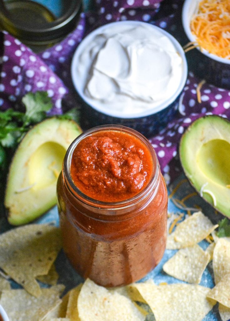 homemade taco sauce in a glass jar surrounded by tortillas chips with two halves of a ripe green a ripe avocado and a bowl of sour cream in the background