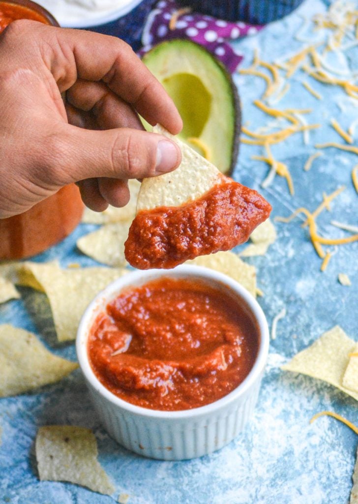 a hand is holding a tortilla chip with homemade taco sauce on it aloft over a white bowl filled with taco sauce on a light blue background