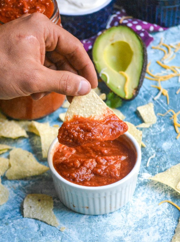 a hand is holding a tortilla chip with homemade taco sauce on it aloft over a white bowl filled with taco sauce on a light blue background