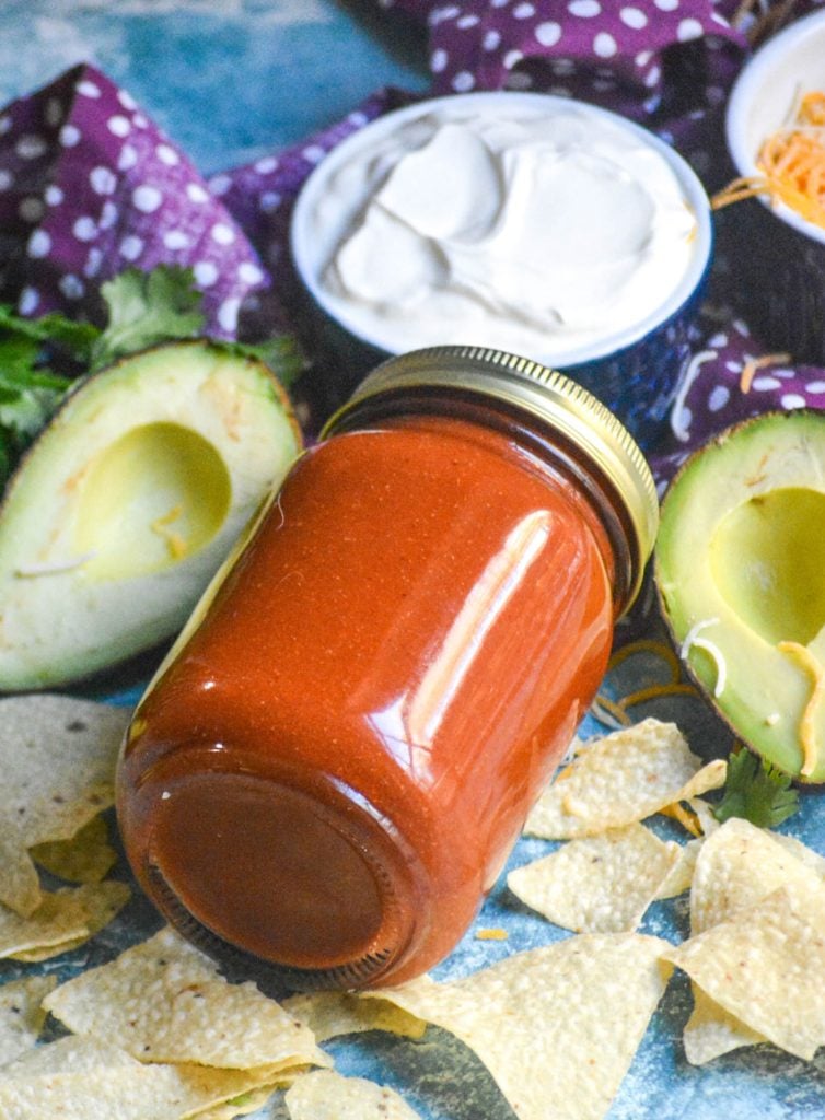 taco sauce in a sealed glass jar laid on it's side and surrounded by chips, a sliced open avocado and a blue bowl filled with fresh sour cream