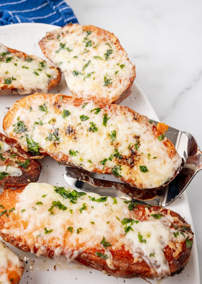 a silver spatula lifting a baked cheesy garlic roasted sweet potato off a white serving tray