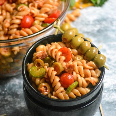 bloody mary pasta salad is shown in a small black serving bowl, stacked atop another empty black bowl with a toothpick strung with green Spanish olives as a garnish