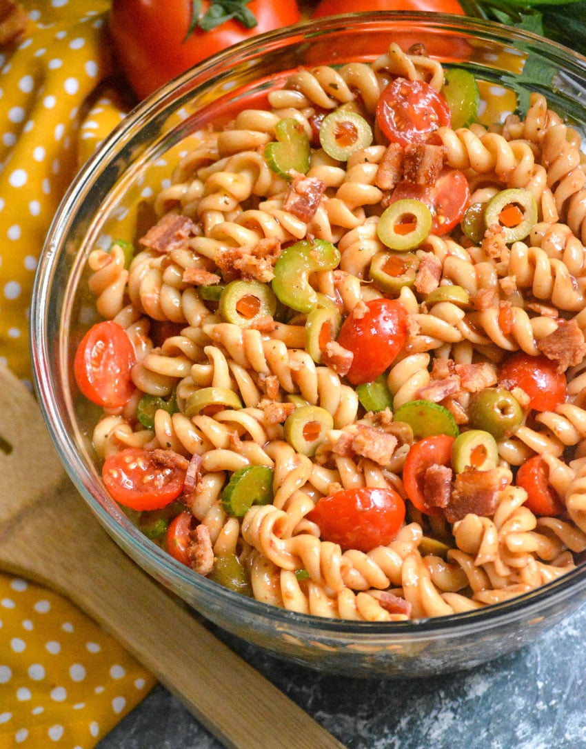 Bloody mary pasta salad in a glass serving bowl on a polka dotted yellow napkin with a wooden spoon for serving