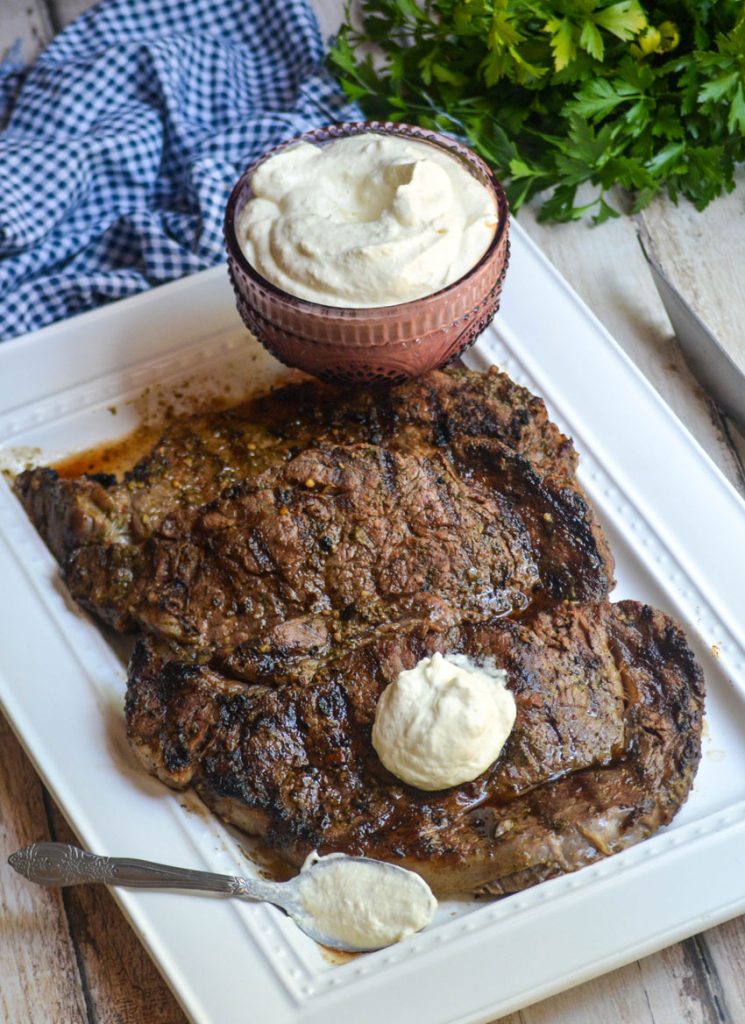 creamy horseradish sauce in a purple glass serving bowl shown with grilled steaks on a white platter topped with a dollop of sauce