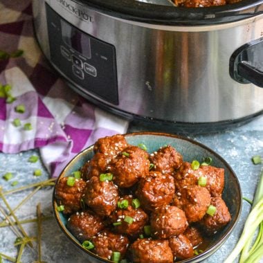 crockpot Asian meatballs shown in a bowl topped with sesame seeds & sliced green onions with the rest in a slow cooker in the background
