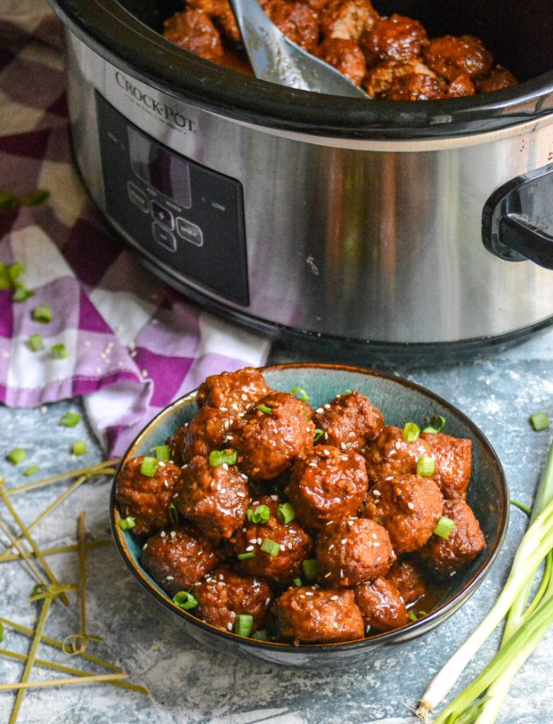 crockpot Asian meatballs shown in a bowl topped with sesame seeds & sliced green onions with the rest in a slow cooker in the background