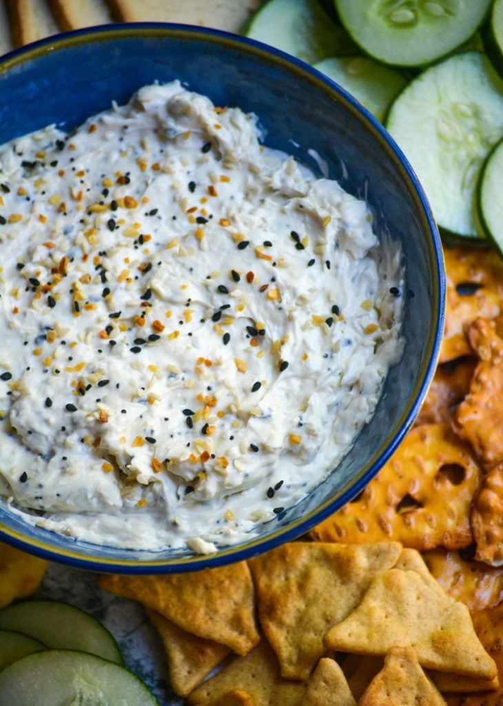 everything bagel dip in blue serving bowl surrounded by crackers, toasts, sliced cucumbers, pretzels, and pita bread