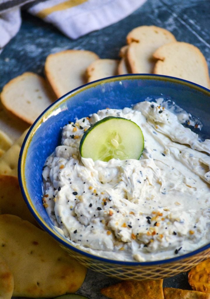 everything bagel dip in a blue serving bowl with a cucumber slice shown being dipped into it