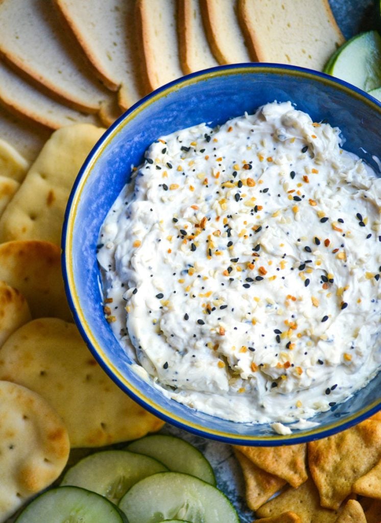 everything bagel dip in blue serving bowl surrounded by crackers, toasts, sliced cucumbers, pretzels, and pita bread
