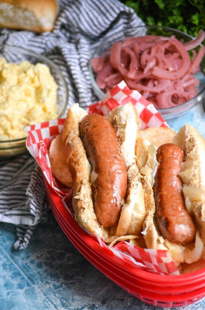 instant pot beer brats stacked in a red picnic basket with pickled onions and potato salad in the background