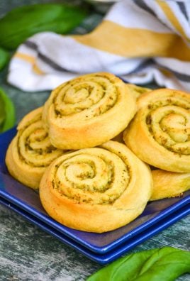 soft buttery pesto lined pinwheels are stacks on a blue appetizer plate with fresh green basil leaves and a striped dishcloth in the back ground