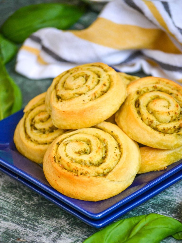 soft buttery pesto lined pinwheels are stacks on a blue appetizer plate with fresh green basil leaves and a striped dishcloth in the back ground