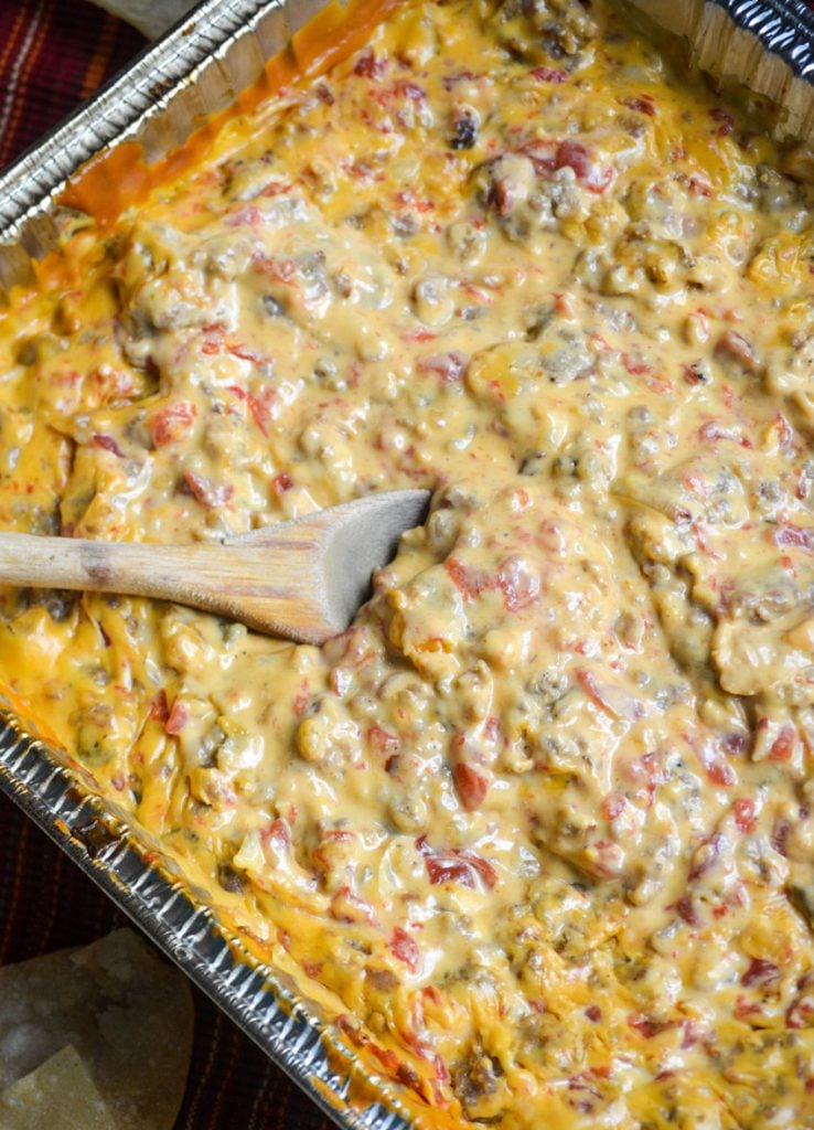 smoked queso dip in an aluminum foil baking pan with a wooden spoon shown dipped in the center