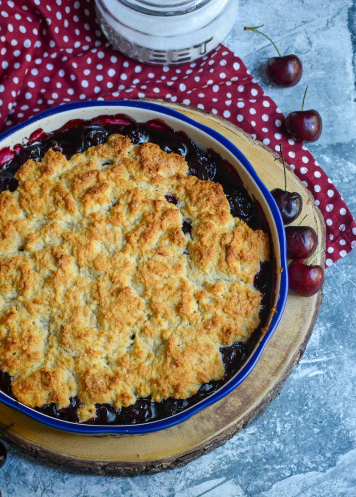 sweet dark cherry cobbler in a blue pie dish on a wooden cutting board with dark cherries and a sugar dish in the background