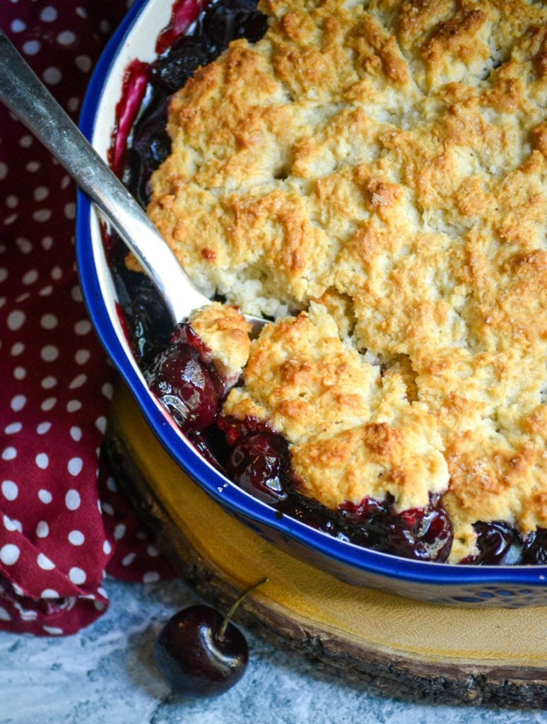 cherry cobbler in a blue ceramic pie dish shown with a silver spoon in it