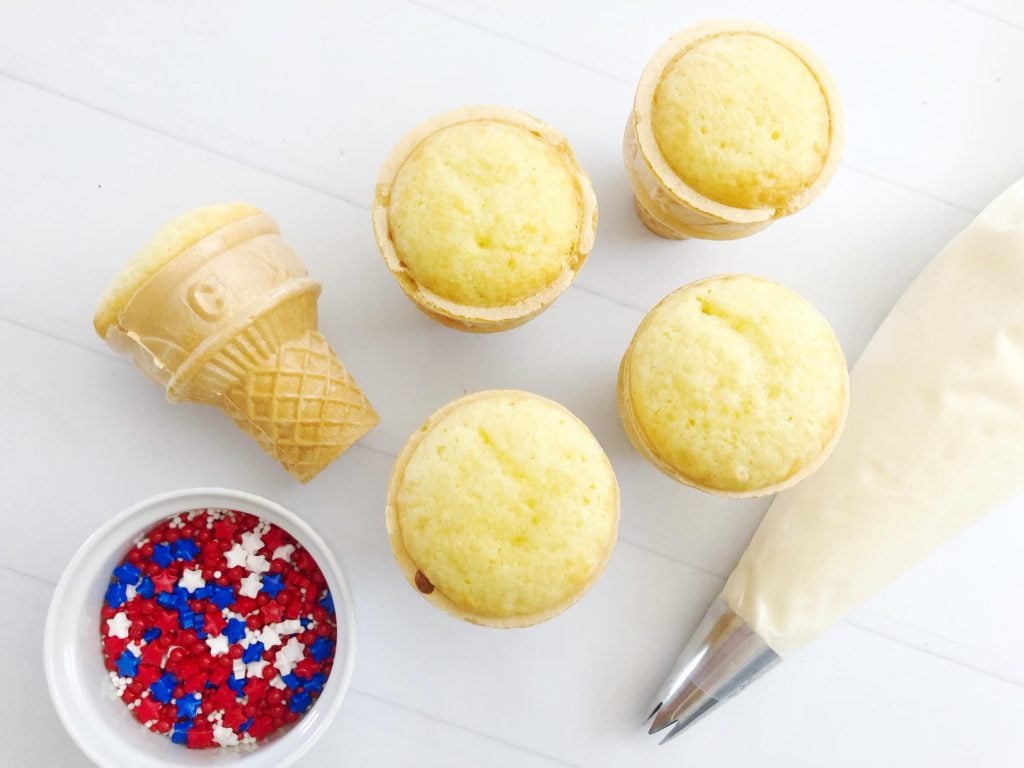 cupcakes baked into sundae cones with a white bowl filled with red white & blue sprinkles and a piping bag filled with white buttercream in the background