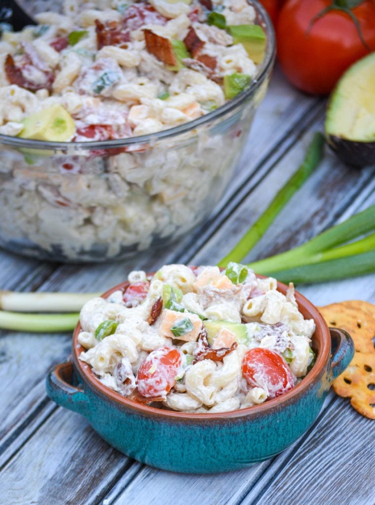 a serving of chicken club pasta salad shown in a blue bowl set in from of fresh produce and the serving bowl