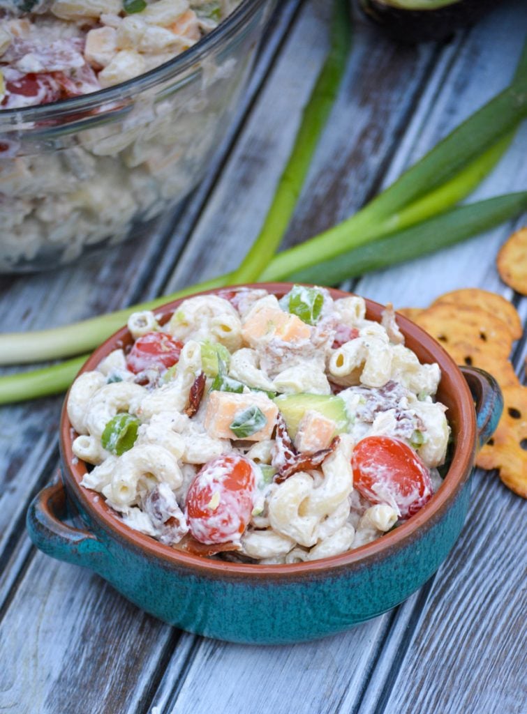a serving of chicken club pasta salad shown in a blue bowl set in from of fresh produce and the serving bowl