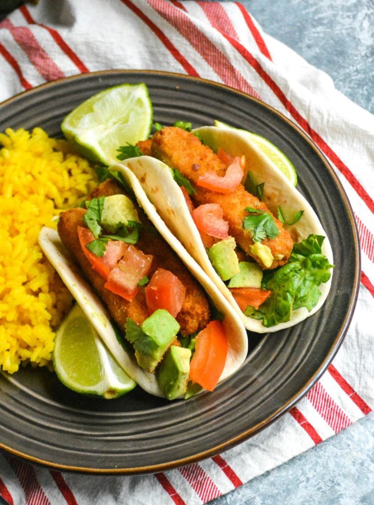 two chili lime fish stick tacos shown on a brown plate with fresh lime wedges and yellow rice