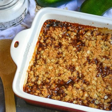 mock apple crisp in a square baking dish shown with a wooden serving spoon, sugar, and ripe zucchini in the background