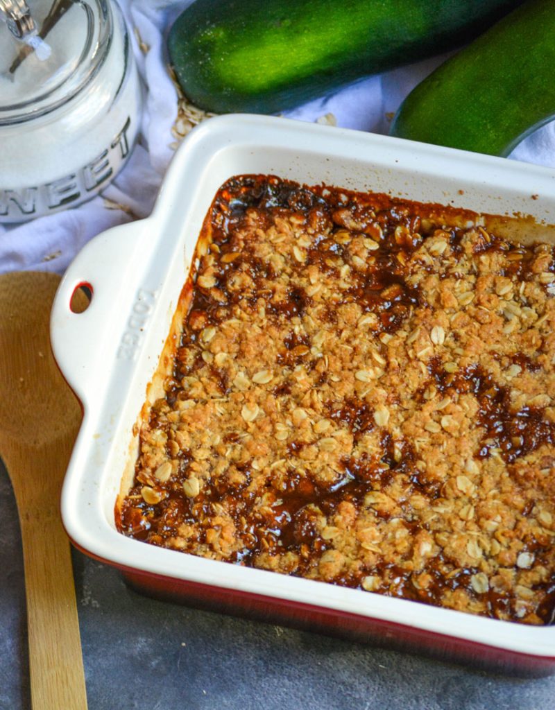 mock apple crisp in a square baking dish shown with a wooden serving spoon, sugar, and ripe zucchini in the background