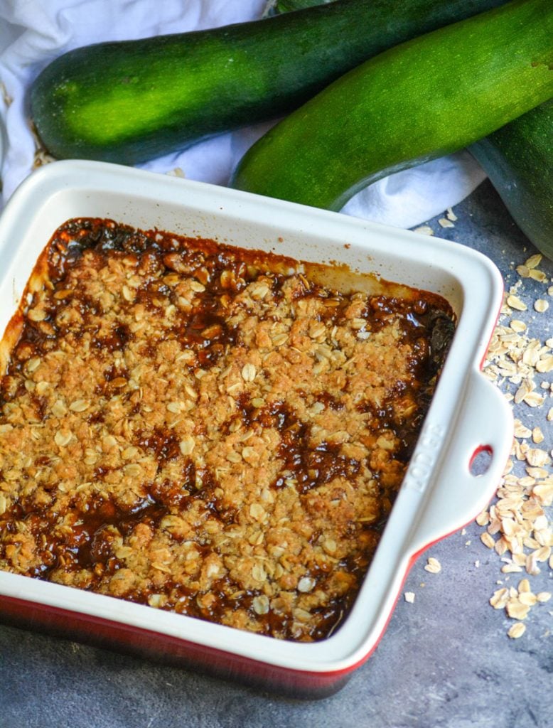 mock apple crisp in a square baking dish shown with a wooden serving spoon, sugar, and ripe zucchini in the background