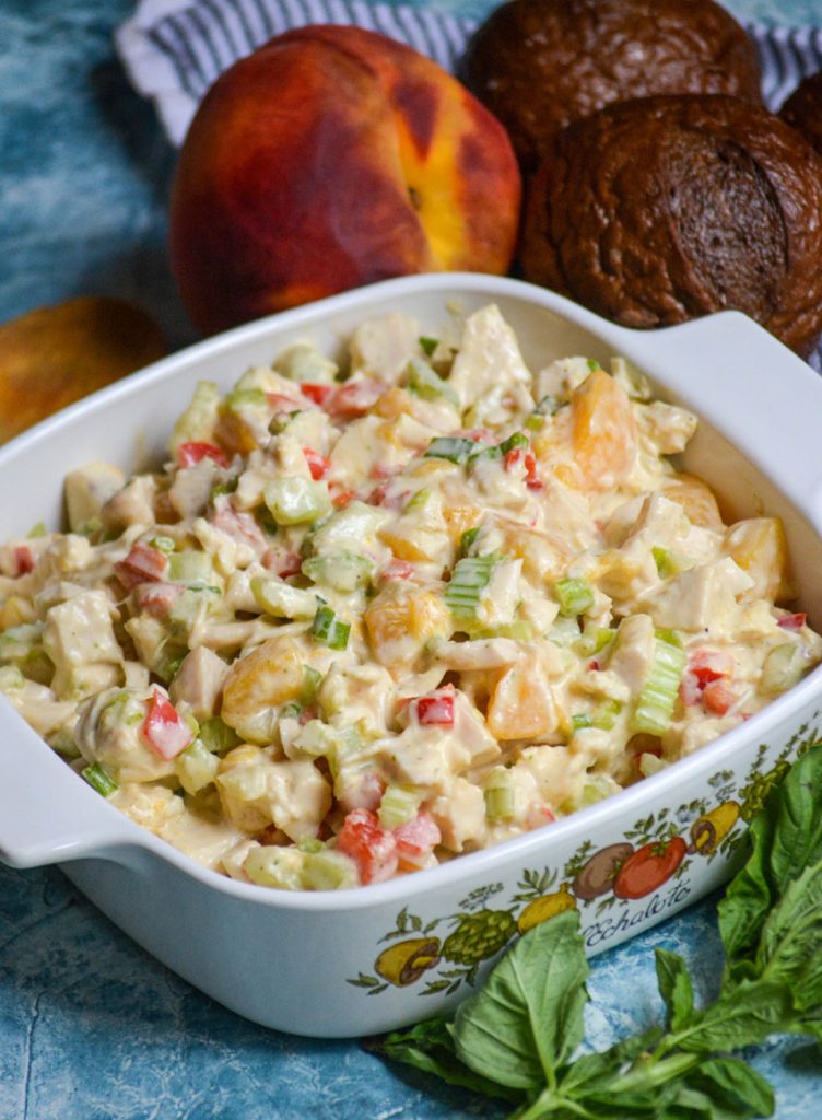 chicken salad with peaches and fresh basil in a white square pyrex dish surrounded by basil leaves, ripe peaches, and pumpernickel rolls