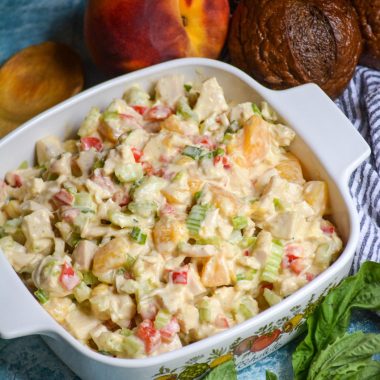 chicken salad with peaches and fresh basil in a white square pyrex dish surrounded by basil leaves, ripe peaches, and pumpernickel rolls