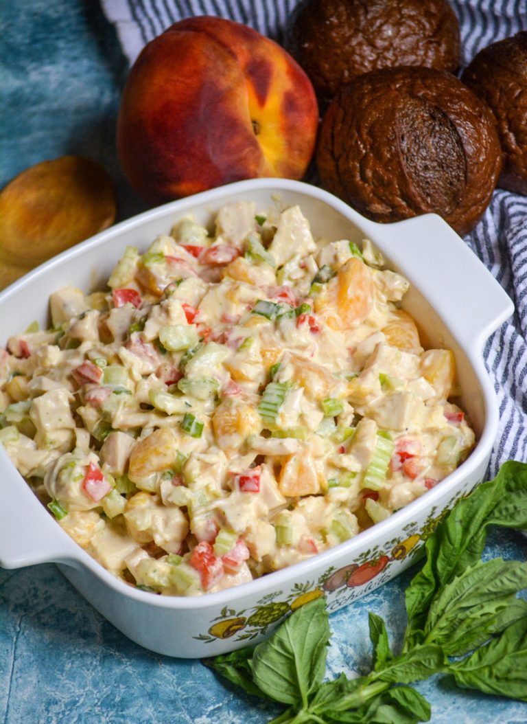 chicken salad with peaches and fresh basil in a white square pyrex dish surrounded by basil leaves, ripe peaches, and pumpernickel rolls