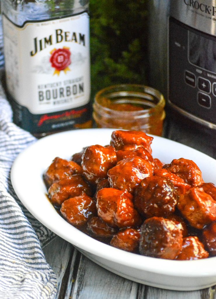 peach bourbon meatballs served in a white dish with a bottle of bourbon, peach syrup, and crockpot in the background